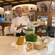 Chef at a table in a restaurant displaying ingredients such as cheese, basil, pine nuts and olive oil, holding a plate of pasta
